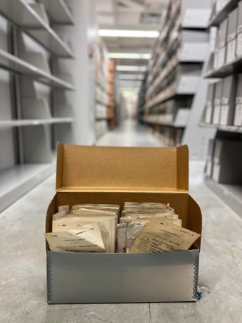 archival box sitting on the floor in between shelves of paper records. Lid to box is open and paper contents visible.