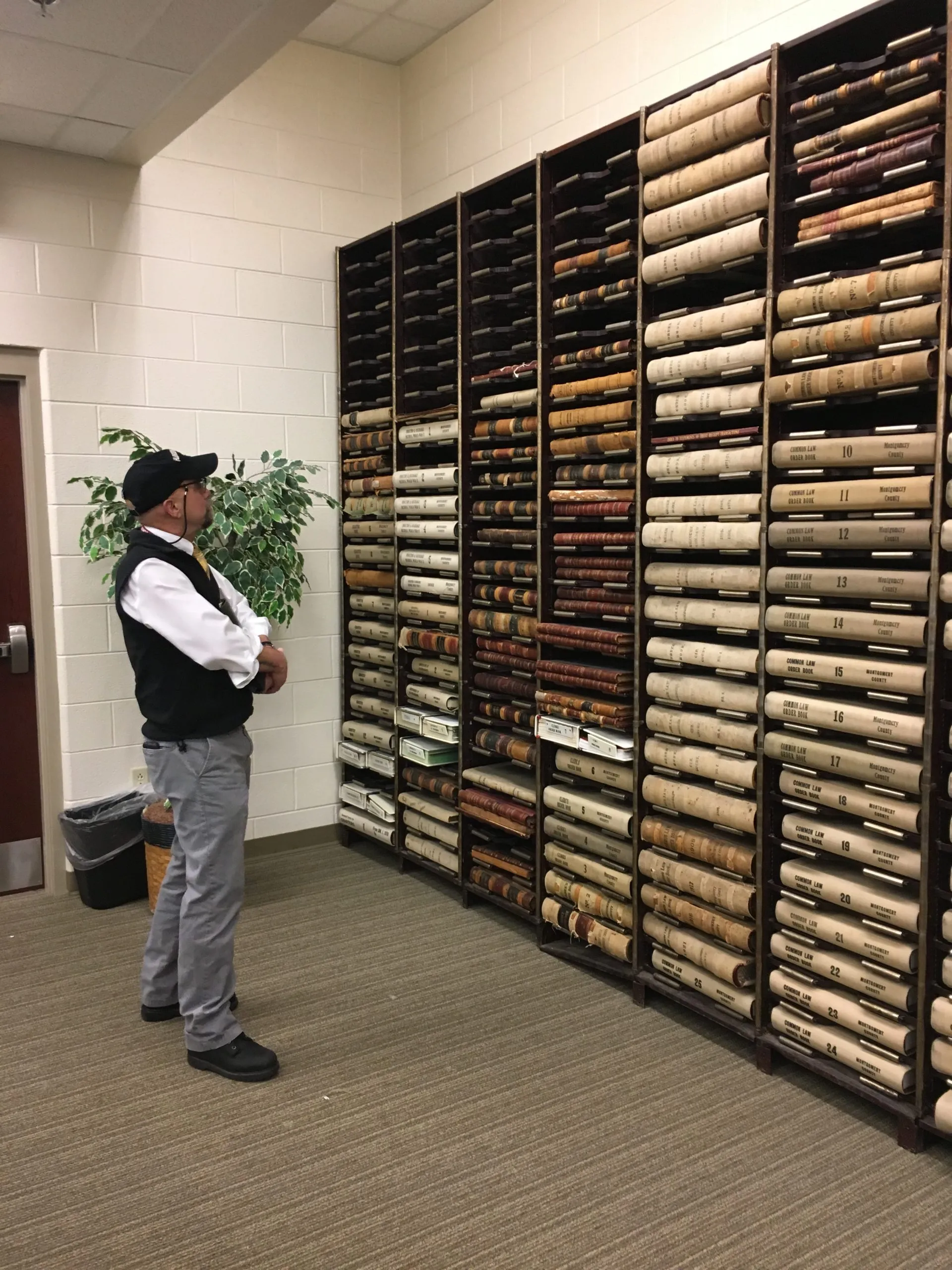 man with baseball hat looking at bound volumes on shelving unit against wall. Volumes are laying on their sides spines facing out.