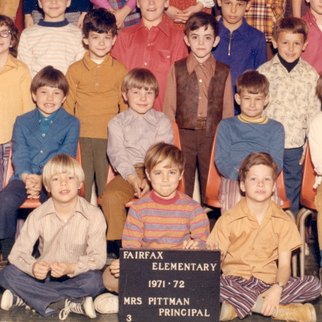 The image is a sample from a school class picture. Three boys sit cross-legged in the front, holding a sign that says Fairfax Elementary 1971-72 Mrs. Pittman Principal" with a "3" in the bottom left, possibly indicating the students being in 3rd grade. There are 3 male students seated behind them, and there are more students standing behind that row.